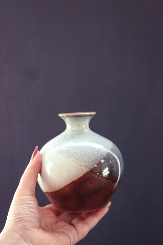 May include: A small, round ceramic vase with a white and brown glaze. The vase has a narrow neck and a wide, rounded body. The glaze is uneven, creating a mottled effect.
